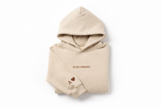 Custom couples embroidered beige hoodie with brown thread by Sharp Stitch, featuring personalized roman numerals on the chest and initials with a heart on the sleeve. Professional custom embroidery and DTF printing for hoodies and crewneck sweatshirts, casual wear, workwear, and uniforms, with nationwide shipping across the USA.