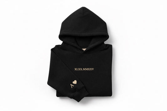 Black couples hoodie with tan thread custom embroidery by Sharp Stitch, showing roman numeral embroidery on the chest and initial with heart detail on the sleeve. Custom embroidered hoodies and crewneck sweatshirts for casual wear, work uniforms, and everyday apparel, shipped nationwide across the United States.