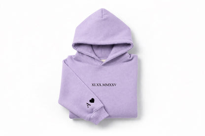 Lavender couples embroidered hoodie with black thread by Sharp Stitch, featuring custom roman numeral embroidery on the chest and initial and heart detail on the sleeve. Premium custom embroidery for hoodies and crewneck sweatshirts, perfect for casual outfits, workwear, and uniforms, with nationwide USA shipping.