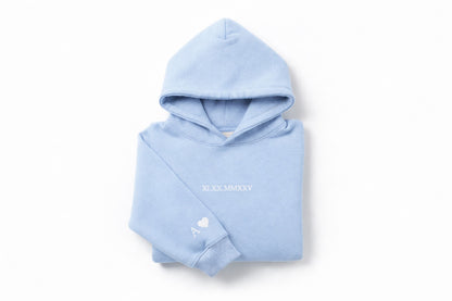Light blue couples embroidered hoodie with white thread by Sharp Stitch, showcasing personalized roman numerals on the chest and initial with heart embroidery on the sleeve. Custom embroidery and DTF printing on hoodies and crewneck sweatshirts for casual wear and workwear, available with nationwide shipping across the United States.