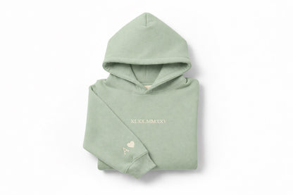 Light green hoodie with tan thread couples embroidery by Sharp Stitch, personalized with roman numerals on the chest and initials with heart embroidery on the sleeve. High quality custom embroidery and DTF printing available on hoodies and crewneck sweatshirts for casual wear and workwear, shipping across all US states.