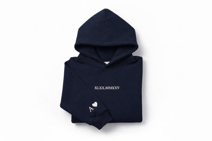 Navy blue couples hoodie with tan thread custom embroidery by Sharp Stitch, designed with roman numerals on the chest and initials with heart embroidery on the sleeve. Professional embroidery and DTF printing on hoodies and crewneck sweatshirts for casual wear, work uniforms, and everyday apparel, shipped nationwide in the USA.
