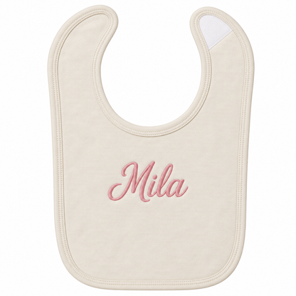 An off-white baby bib with the name 'Mila' embroidered in pink cursive script in the center, shown on a plain white background.

